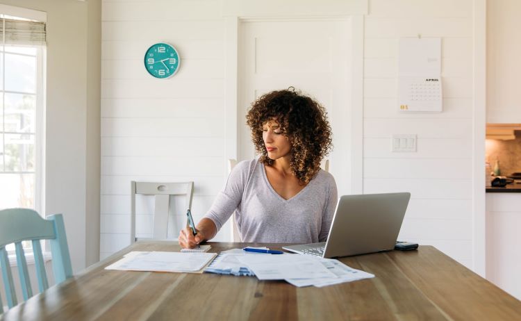 Image of woman working at table in front of laptop and notebooks.