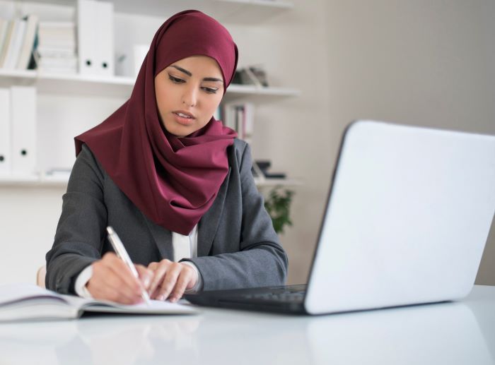 Professional woman at desk with laptop, writing in notebook.