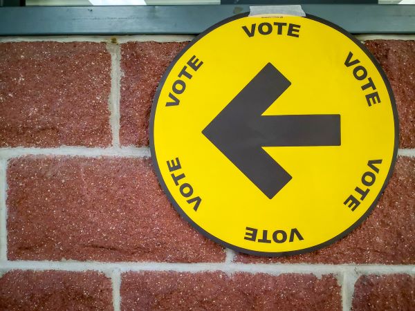 Yellow circular vote sign with arrow on a brick wall.