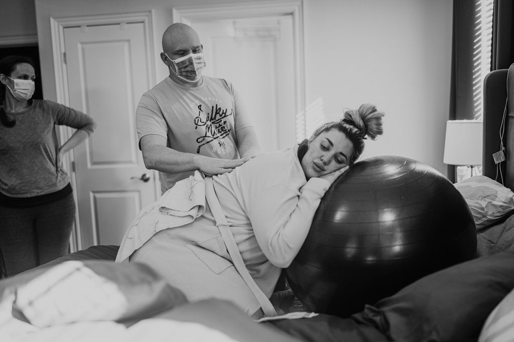 Woman in labour leans on a birth ball on her bed at home while her partner and a midwife stand nearby.