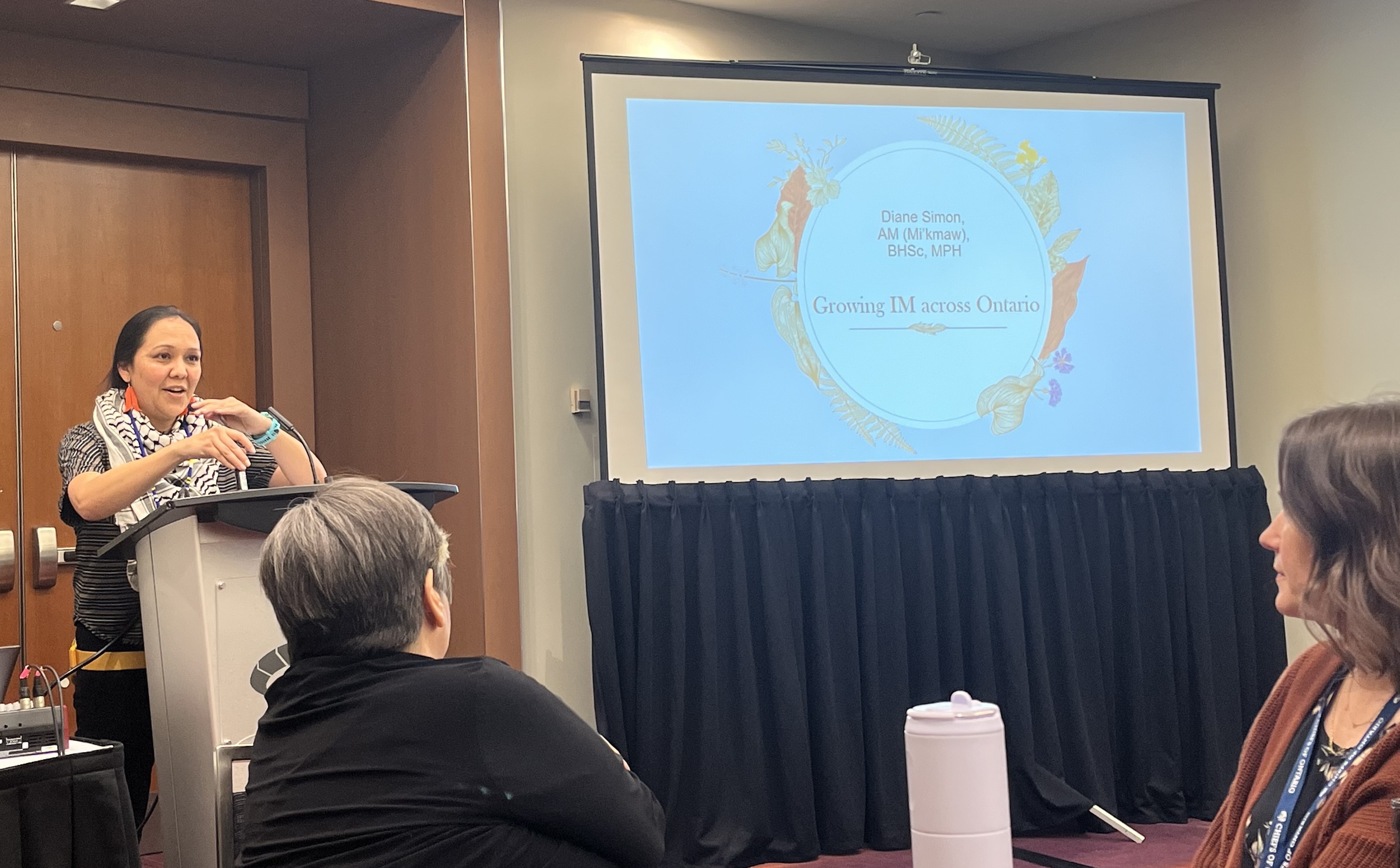 Indigenous Midwife Diane Simon stands at a podium next to a projector screen in front of a captive audience.