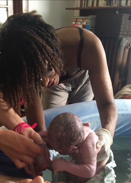 Person with dark curly hair leaning over a birthing tub holding a baby