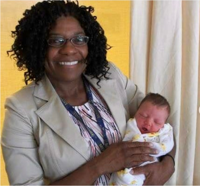 Person with dark curly hair, smiling and wearing glasess, facing the camera, holding a young baby