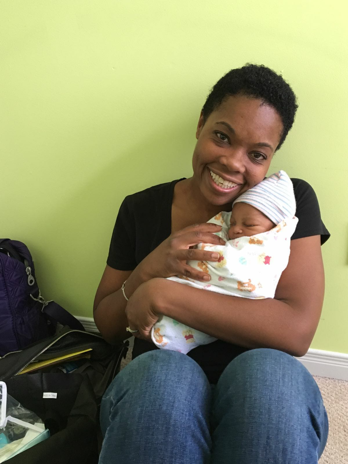 Person sitting against the wall, with short black hair, holding a baby and smiling