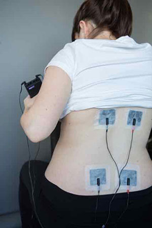 Picture of person’s lower back with four square sticky pads attached to it. The pads are attached to wires which connect the pads to a small, plastic, black box in the person’s hand by wires.
