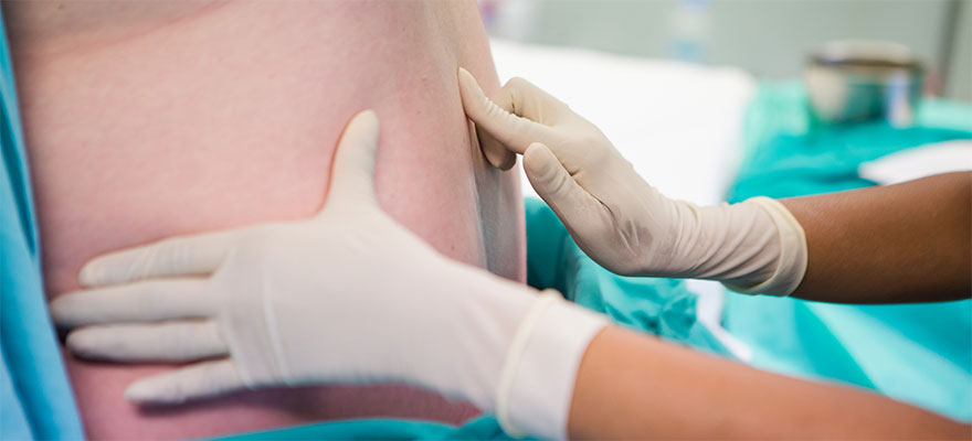 Picture of someone’s lower back with two gloved hands touching it in a hospital bed.