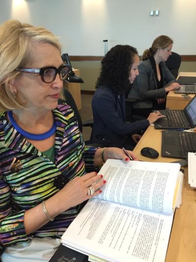 Three women in professional attire seated at a desk with laptops and a large book